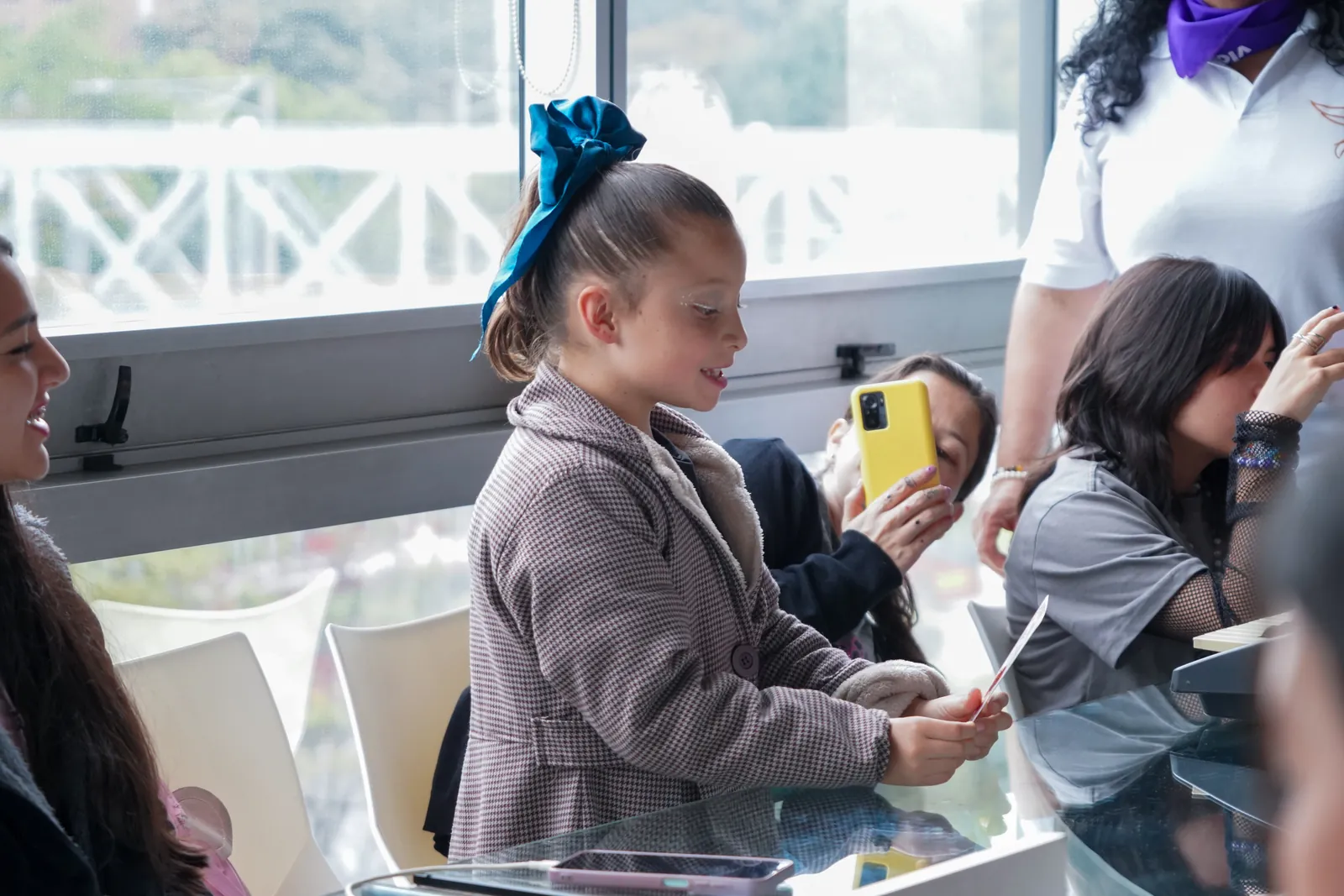 A girl with a blue bow smiles as she reads a card at the table.